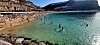 Turister bader og soler seg på en travel sandstrand ved klart havvann omgitt av klipper.