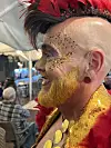 Person med gul skjegg, glitter og fjærkostyme sitter blant publikum på utekafé.