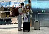 To reisende med munnbind står ved en koffert og bagasjevogn i en flyplassterminal.
